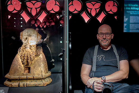 Ein Mann sitzt in der Ausstellung neben einer Vitrine in der sich eine Skulptur befindet